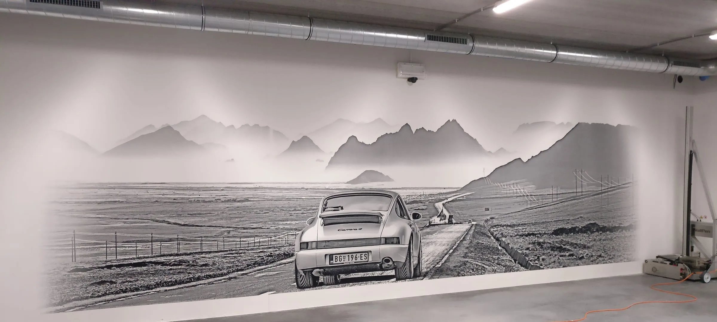 Black and white photo mural of a classic Porsche 911 on a road through misty mountains, printed along a parking garage wall, with the wall printer at the right
