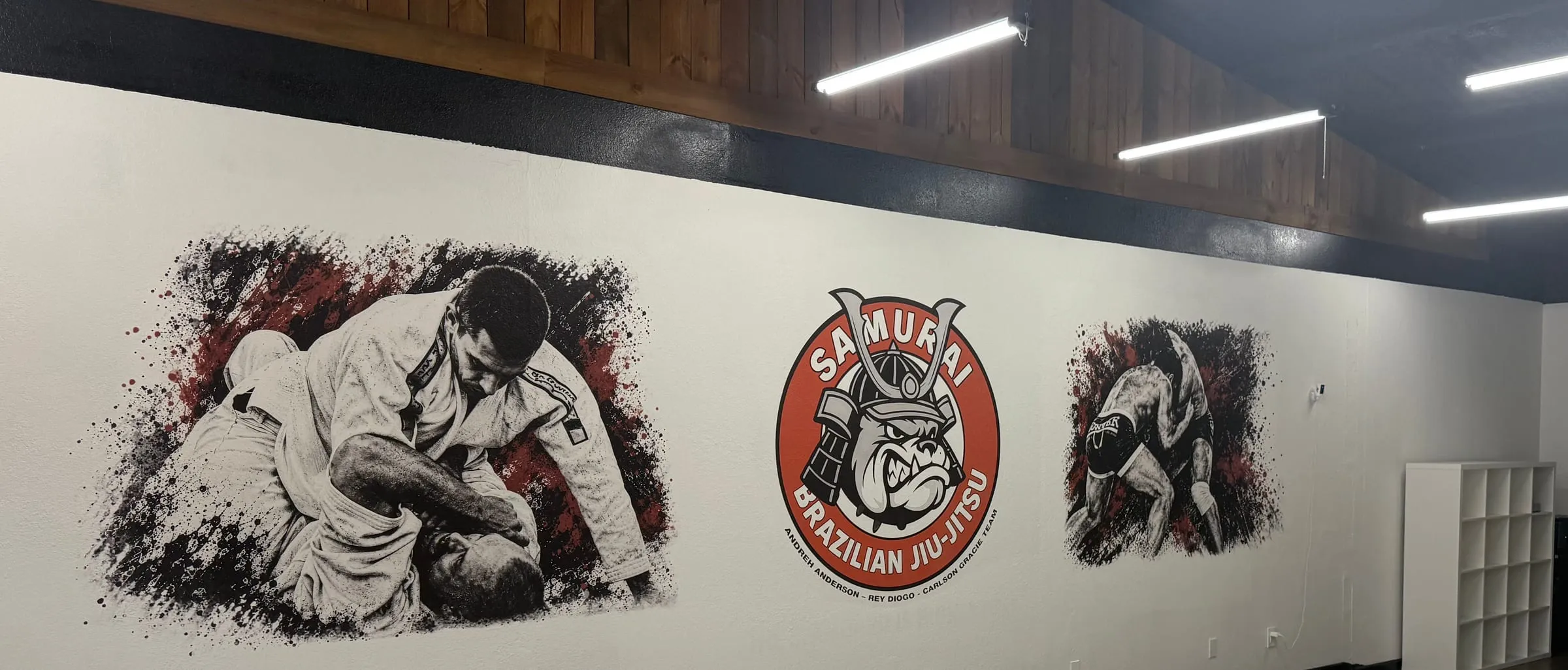 Samurai Brazilian Jiu-Jitsu logo between two black, white, and red photo murals of grapplers on an academy wall